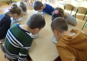Dzieci siedzą przy stoliku jedzą miód z talerzyków bez użycia rąk.